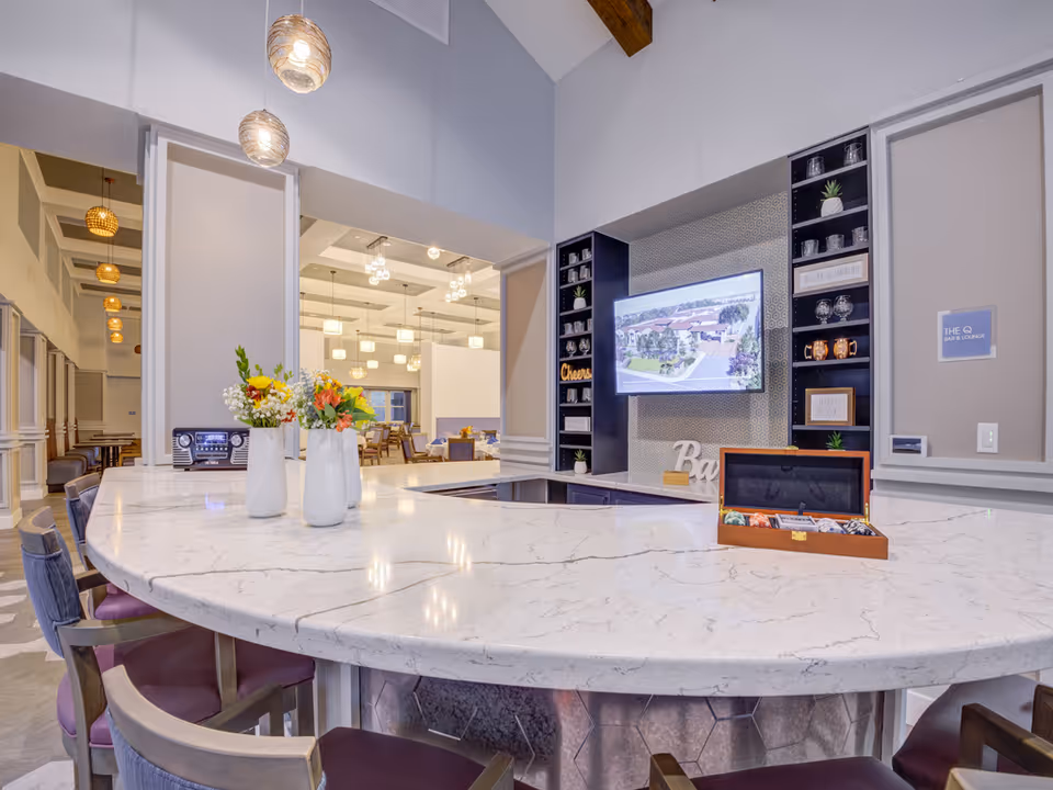 Bright communal bar and lounge area with a curved marble countertop, bar stools, wall-mounted TV, and decorative shelving.