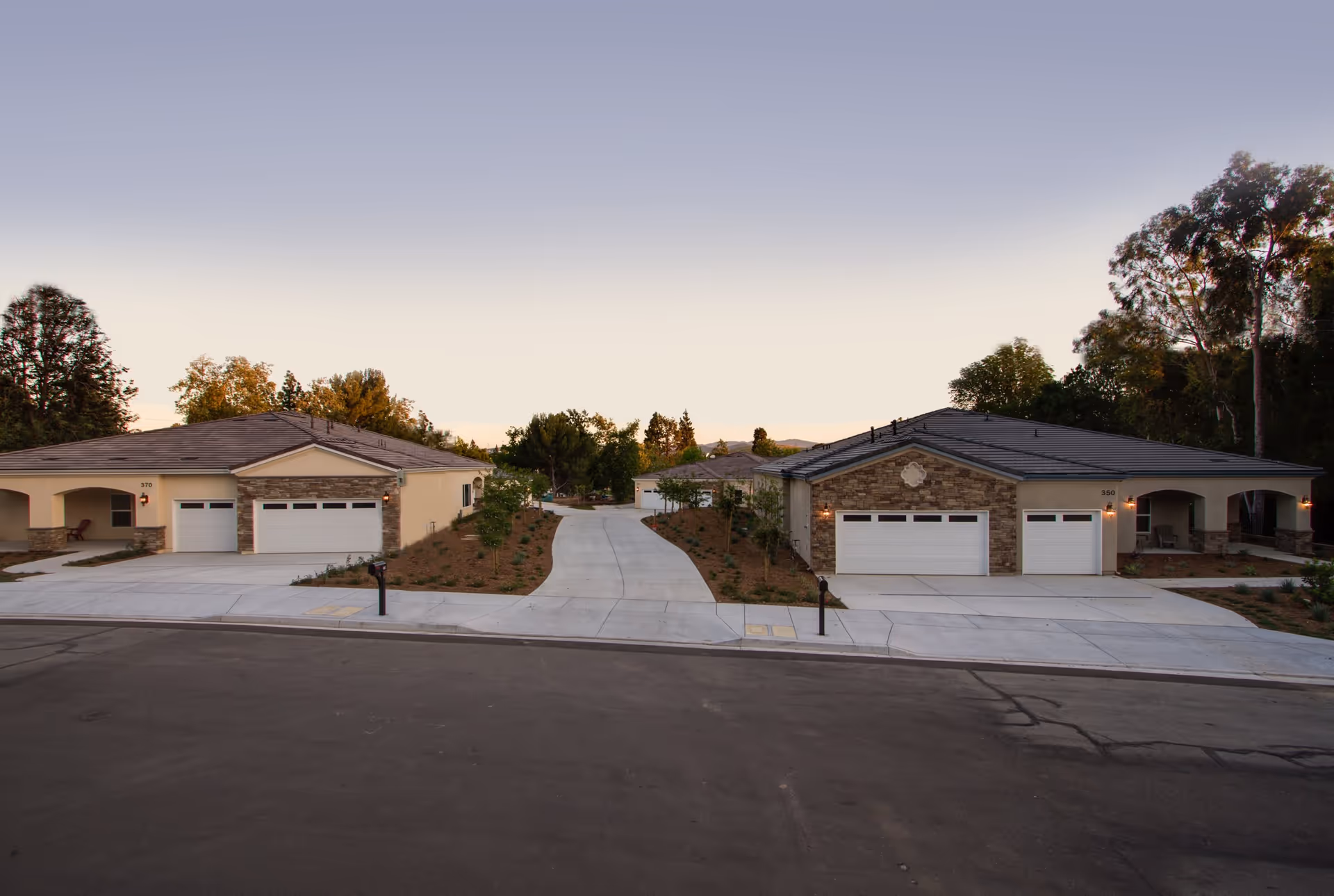 Exterior view of two single-story residential buildings with garages and driveways, surrounded by trees and landscaping under a clear sky at dusk.