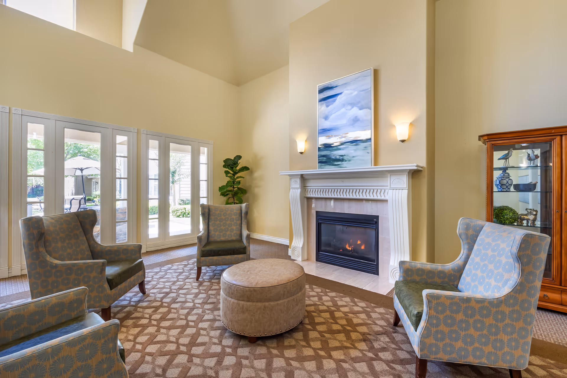A cozy living room area with four patterned armchairs arranged around a round beige ottoman on a patterned carpet. There is a white fireplace with a fire burning and a large abstract painting above it. To the right, there is a wooden display cabinet with decorative items. Large windows and glass doors let in natural light and show an outdoor patio with an umbrella and chairs.