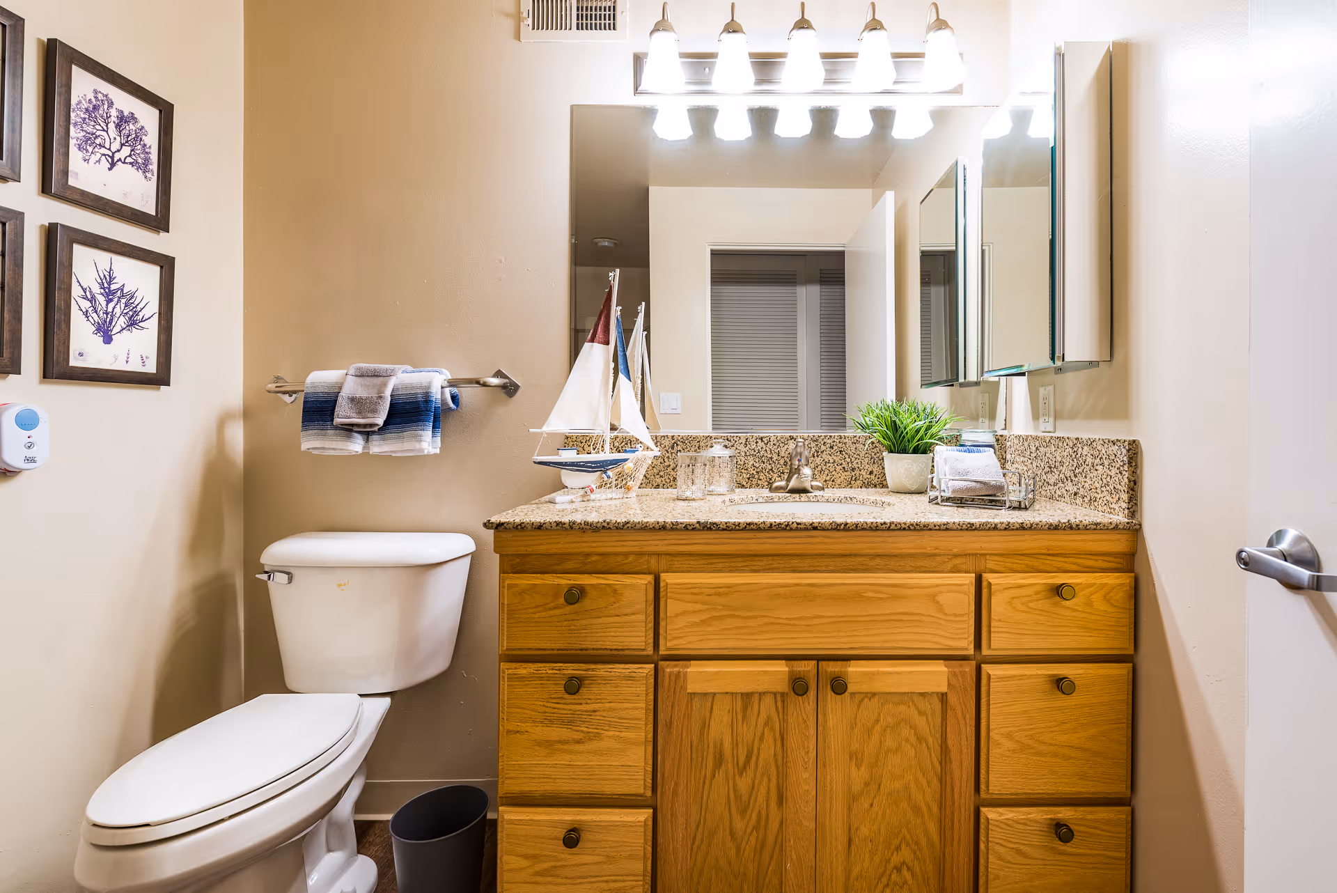 A well-lit bathroom with a wooden vanity and granite countertop, sink and mirror, a toilet, and decorative accents.