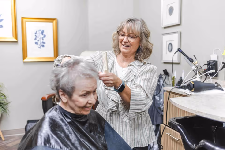 A senior woman with short gray hair is getting her hair styled by a hairstylist in a salon setting. The hairstylist is smiling and wearing glasses and a striped blouse. The room has framed artwork on the walls and salon equipment like a hair dryer on the counter.