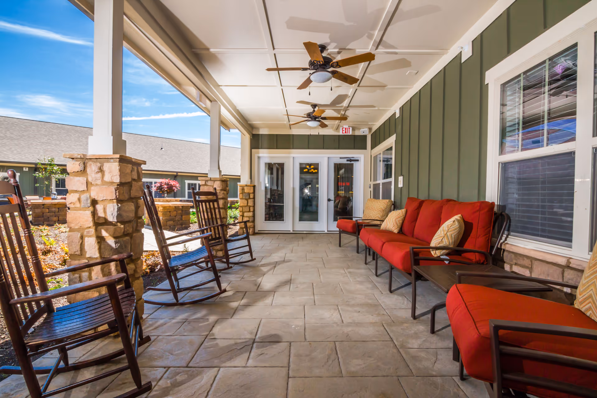 Covered outdoor patio area with stone pillars, ceiling fans, wooden rocking chairs, and red cushioned seating along the wall of a green building with windows and double glass doors.
