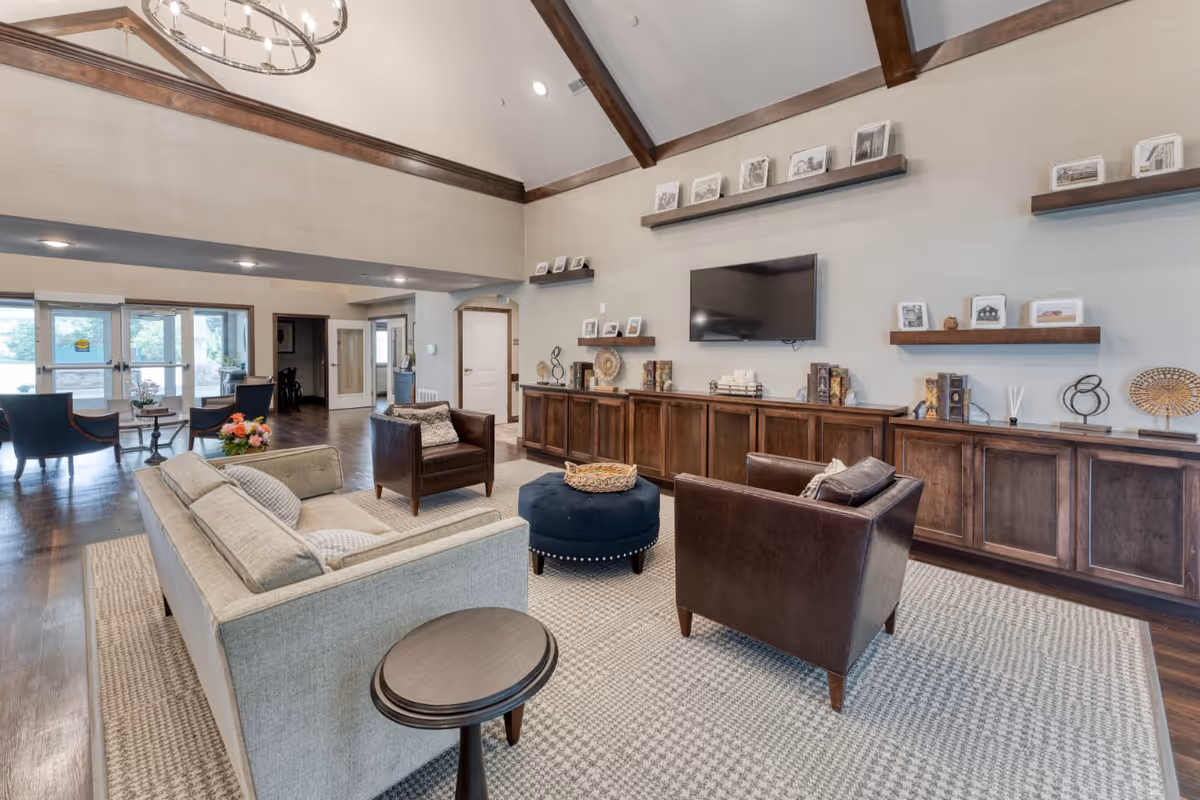 A spacious senior living facility common area with a high vaulted ceiling featuring wooden beams and a modern chandelier. The room includes a beige sofa, two brown leather armchairs, a round blue ottoman, and a small round side table on a patterned rug. The far wall has built-in wooden cabinets with decorative items and framed photos on floating shelves, and a flat-screen TV mounted above. Large windows and glass doors provide natural light and a view of the outdoor area.