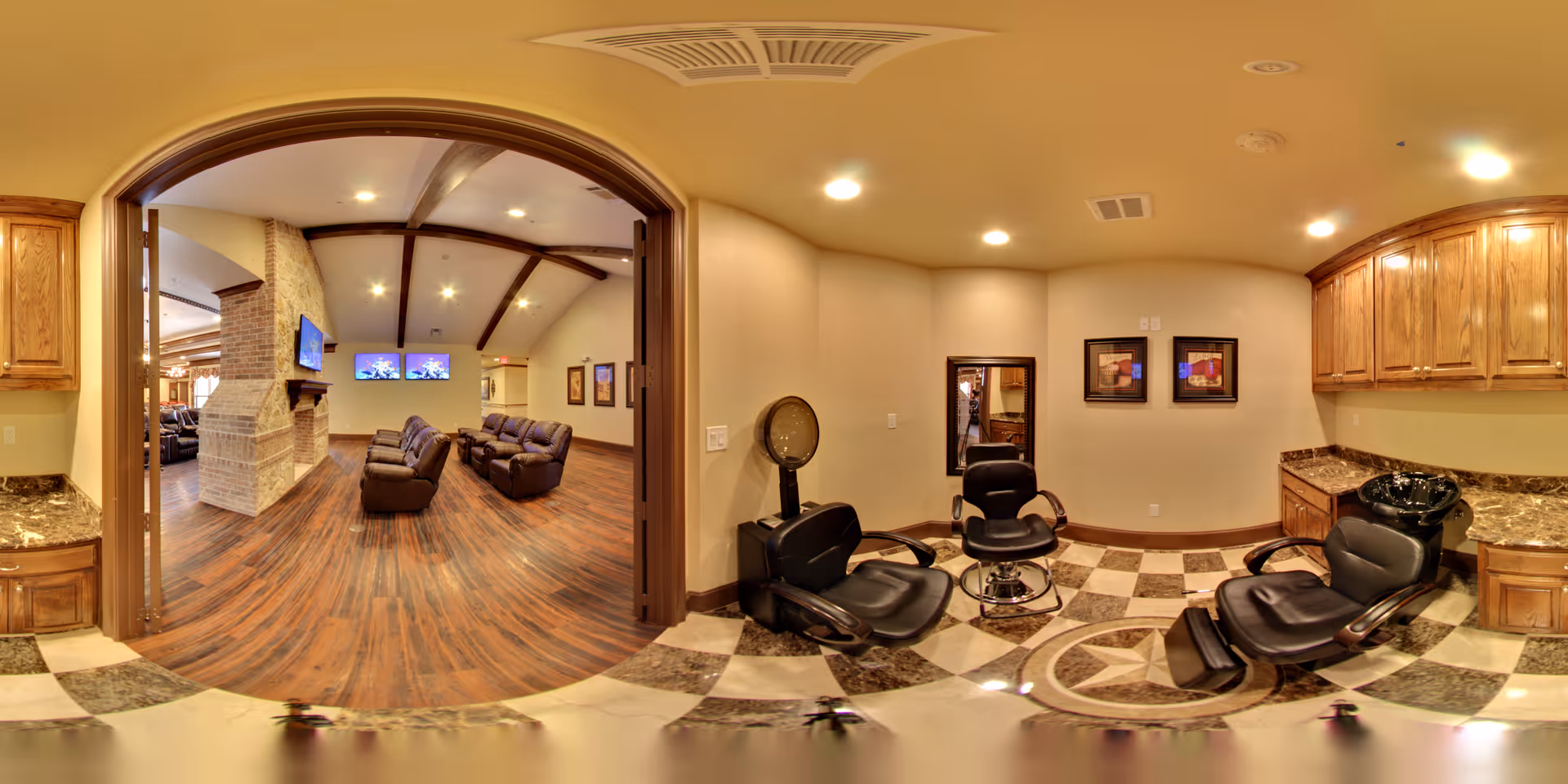 Interior view of a senior living facility showing a hair salon area with three black salon chairs, a hair dryer, and wooden cabinets. Adjacent to the salon is a spacious lounge area with brown leather recliners, a brick fireplace, and two wall-mounted televisions. The floor has a mix of checkered tiles in the salon and wood flooring in the lounge.