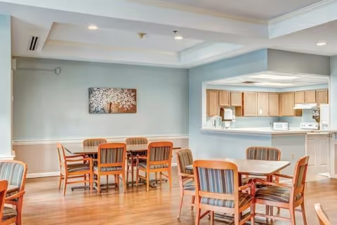 A bright dining area with wooden floors and multiple wooden tables surrounded by chairs with striped upholstery. The room has light blue walls with white trim and a ceiling with recessed lighting. In the background, there is a kitchen area with wooden cabinets, a microwave, and a stove. A piece of artwork featuring a tree with blossoms hangs on the wall.