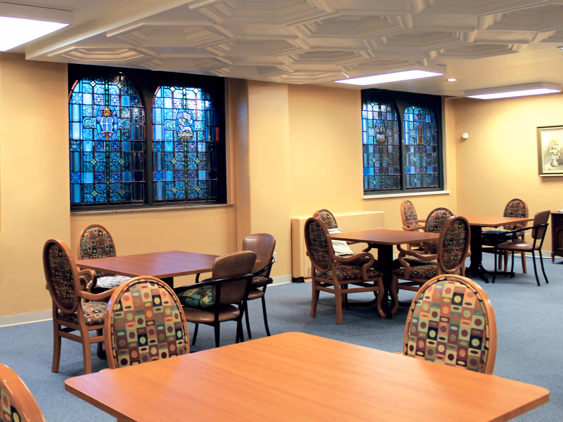 Dining area with multiple wooden tables and patterned upholstered chairs beneath stained glass windows.