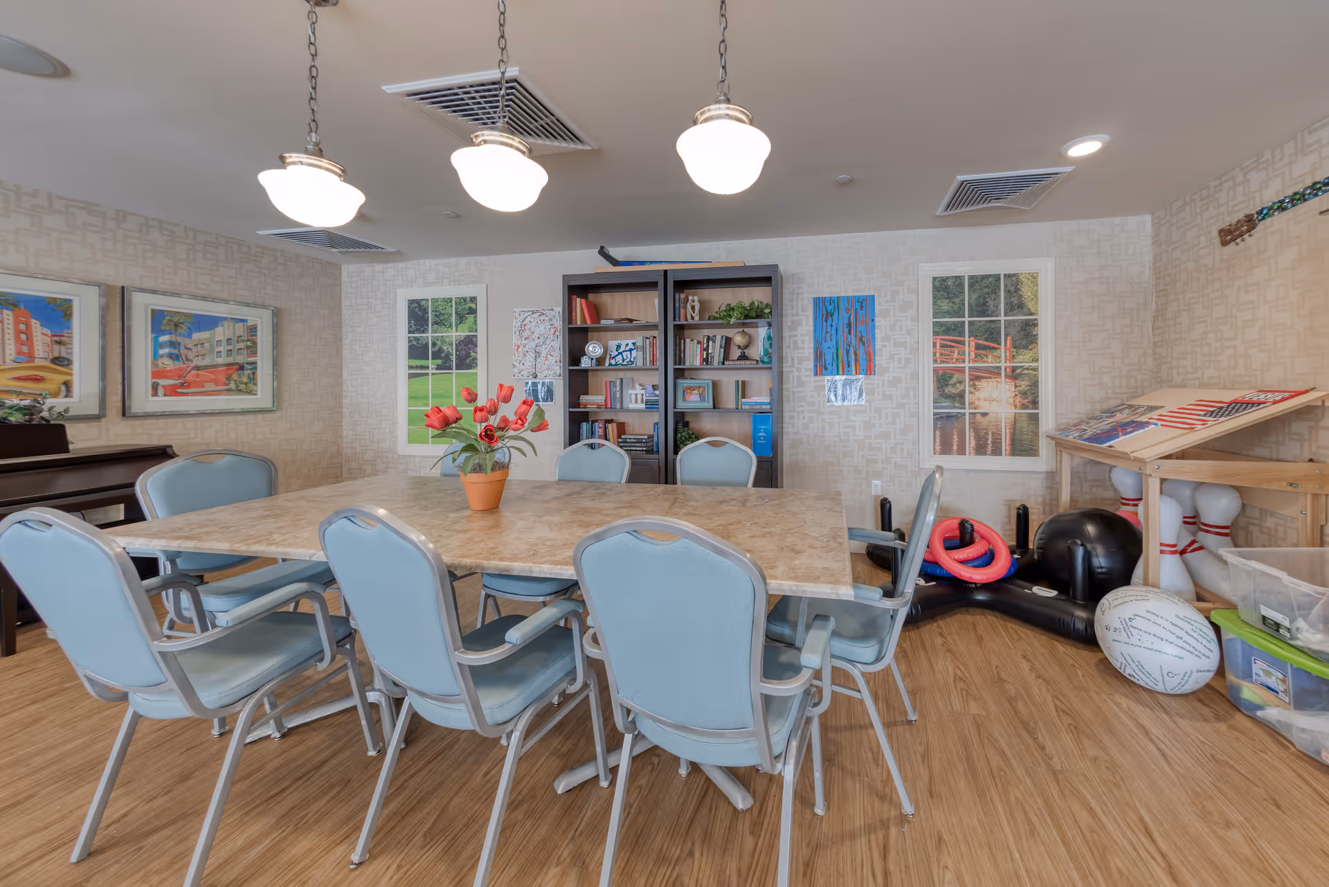 A well-lit room with a rectangular table surrounded by eight light blue cushioned chairs. On the table is a small potted plant with red flowers. The walls have framed pictures and two window-like wall decorations showing outdoor scenes. There is a bookshelf filled with books and decorative items against the back wall. To the right, there is a wooden rack holding bowling pins and balls, along with other recreational equipment. The floor is wood-patterned, and three pendant lights hang from the ceiling above the table.