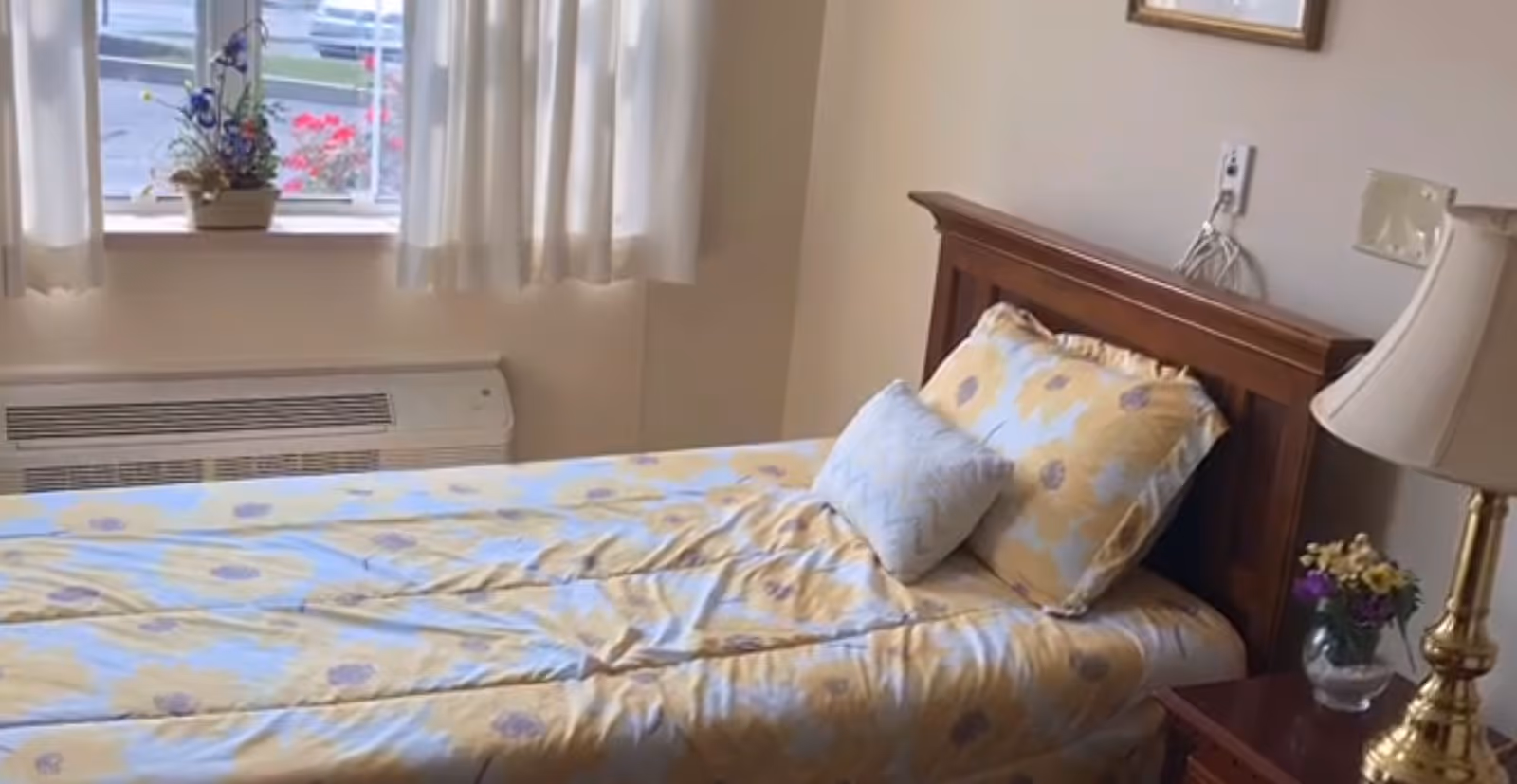 A single bed with a wooden headboard, covered with a yellow and white floral patterned bedspread and two pillows. Next to the bed is a wooden nightstand with a brass lamp and a small vase of flowers. A window with white curtains and a potted plant on the windowsill is visible in the background.