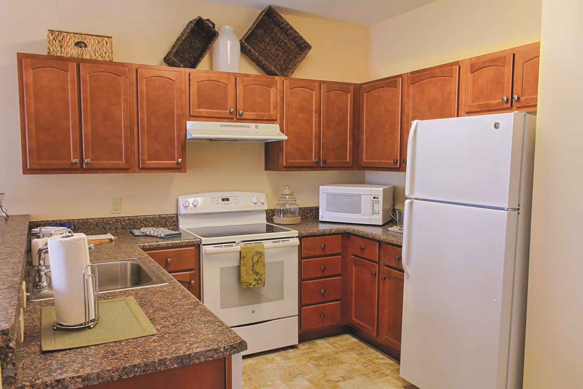 A kitchen with brown wooden cabinets, a white electric stove with a green towel hanging on the handle, a white microwave, a white refrigerator, a stainless steel sink, and a paper towel holder on the counter. There are decorative baskets and a white vase on top of the cabinets.