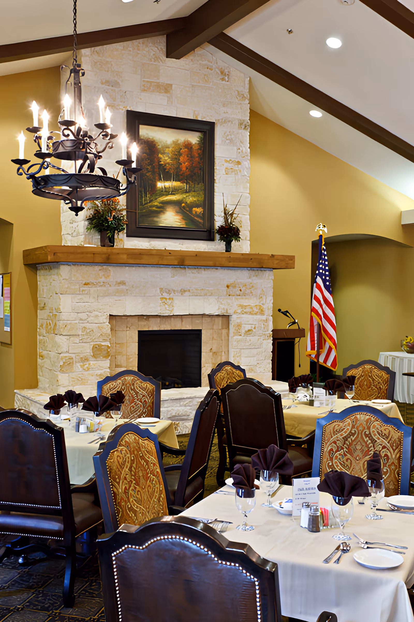 A dining room with several tables set with white tablecloths, glassware, silverware, and folded dark napkins. The room features a large stone fireplace with a wooden mantle, above which hangs a framed painting of a forest path. There is a decorative chandelier hanging from the ceiling and an American flag standing near a podium in the background.