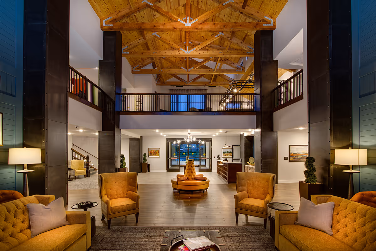 Spacious and warmly lit senior living facility lobby with high wooden beam ceiling, comfortable mustard-colored armchairs and sofas arranged around small tables, a round tufted seating area in the center, and a reception desk in the background.