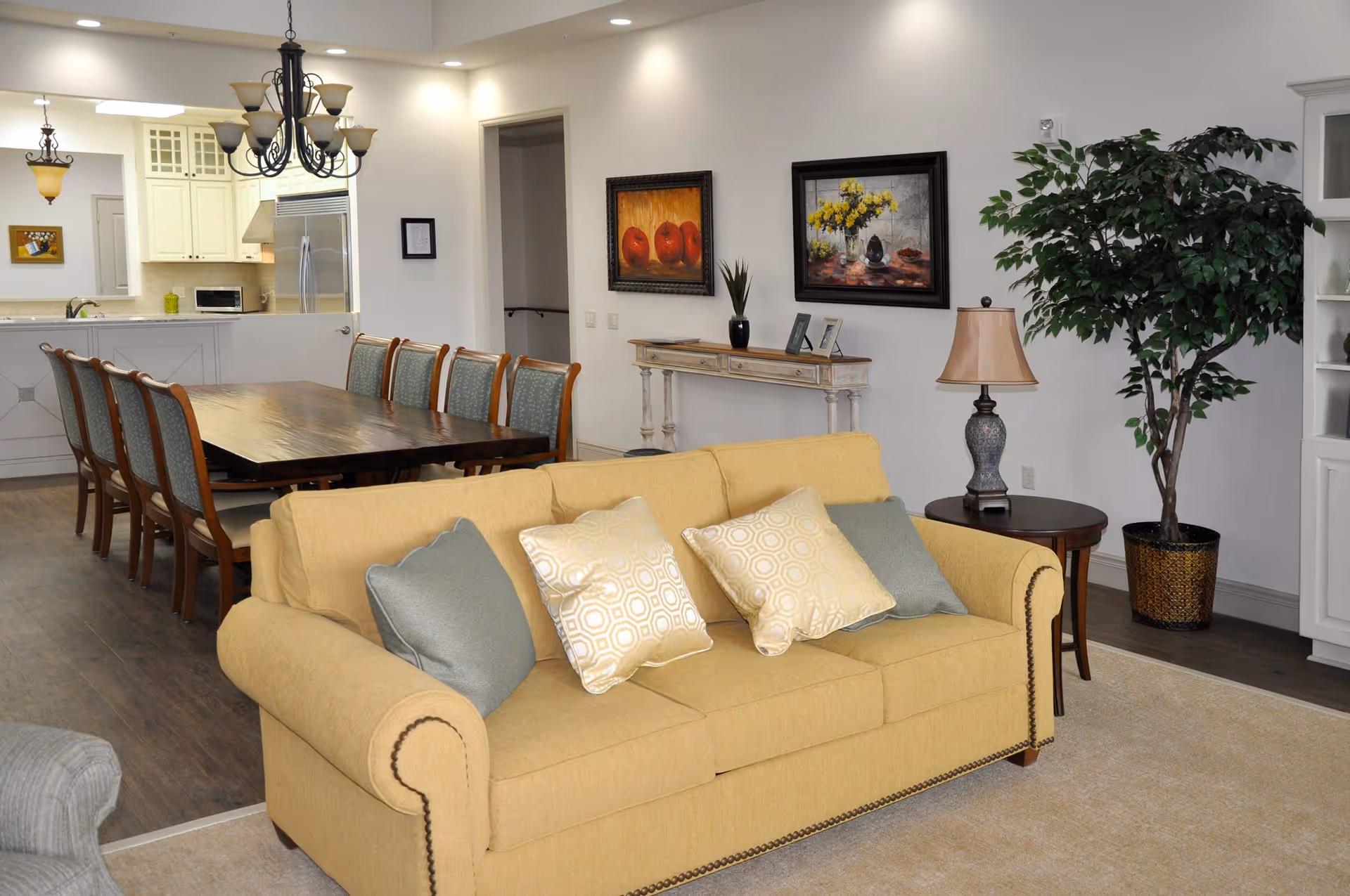 A cozy living room and dining area with a yellow sofa adorned with four decorative pillows, a wooden dining table with eight chairs, a side table with a lamp, a large potted plant, and two framed paintings on the wall. The kitchen is visible in the background with white cabinetry and stainless steel appliances.