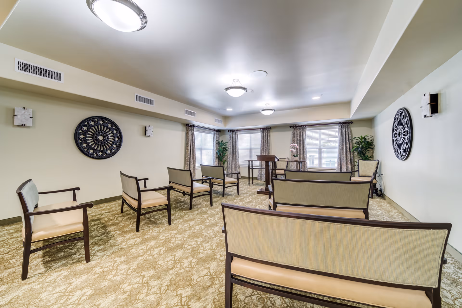 A well-lit room with beige walls and carpet, featuring several cushioned chairs arranged in rows facing a wooden podium and a small table with a flower vase near large windows with curtains. Decorative wall art and plants are placed on the walls and corners.
