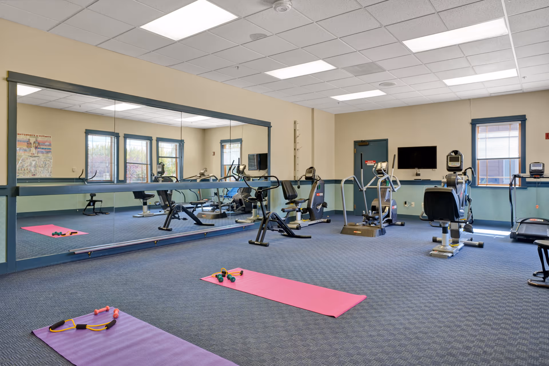 A fitness room with exercise equipment including stationary bikes, elliptical machines, and a treadmill. There are two exercise mats on the carpeted floor with small weights and resistance bands on them. The room has large mirrors on one wall, windows with blue trim, and a wall-mounted television.