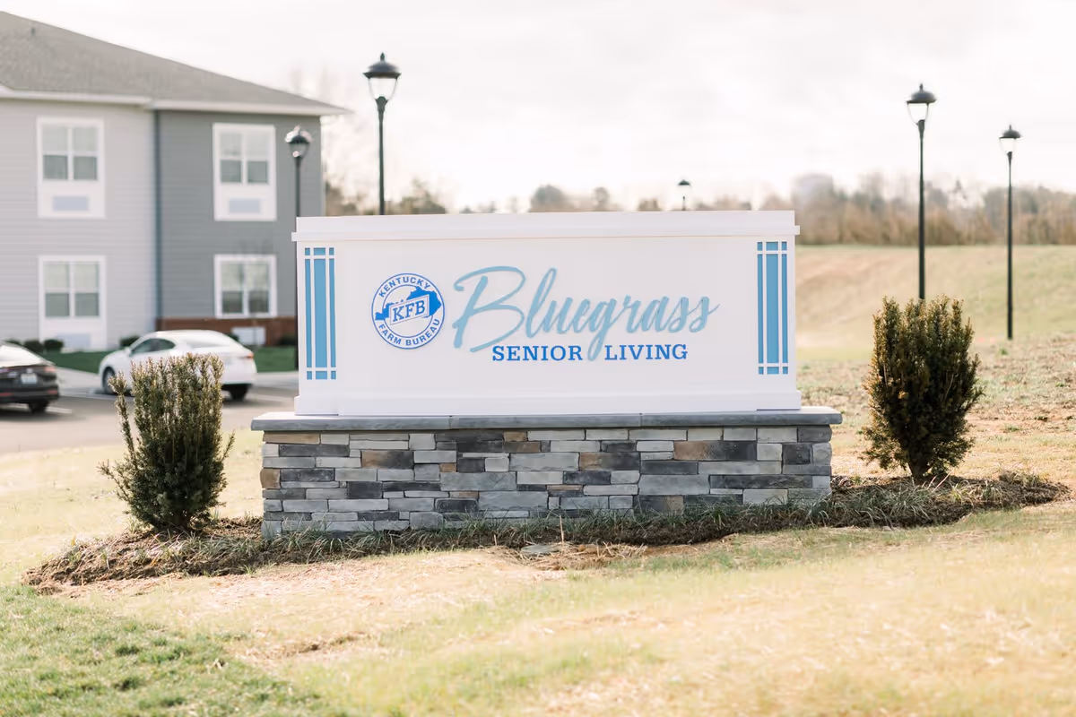 Outdoor sign for Bluegrass Senior Living with the Kentucky Farm Bureau logo, set on a stone base with bushes on either side and a building and street lamps in the background.