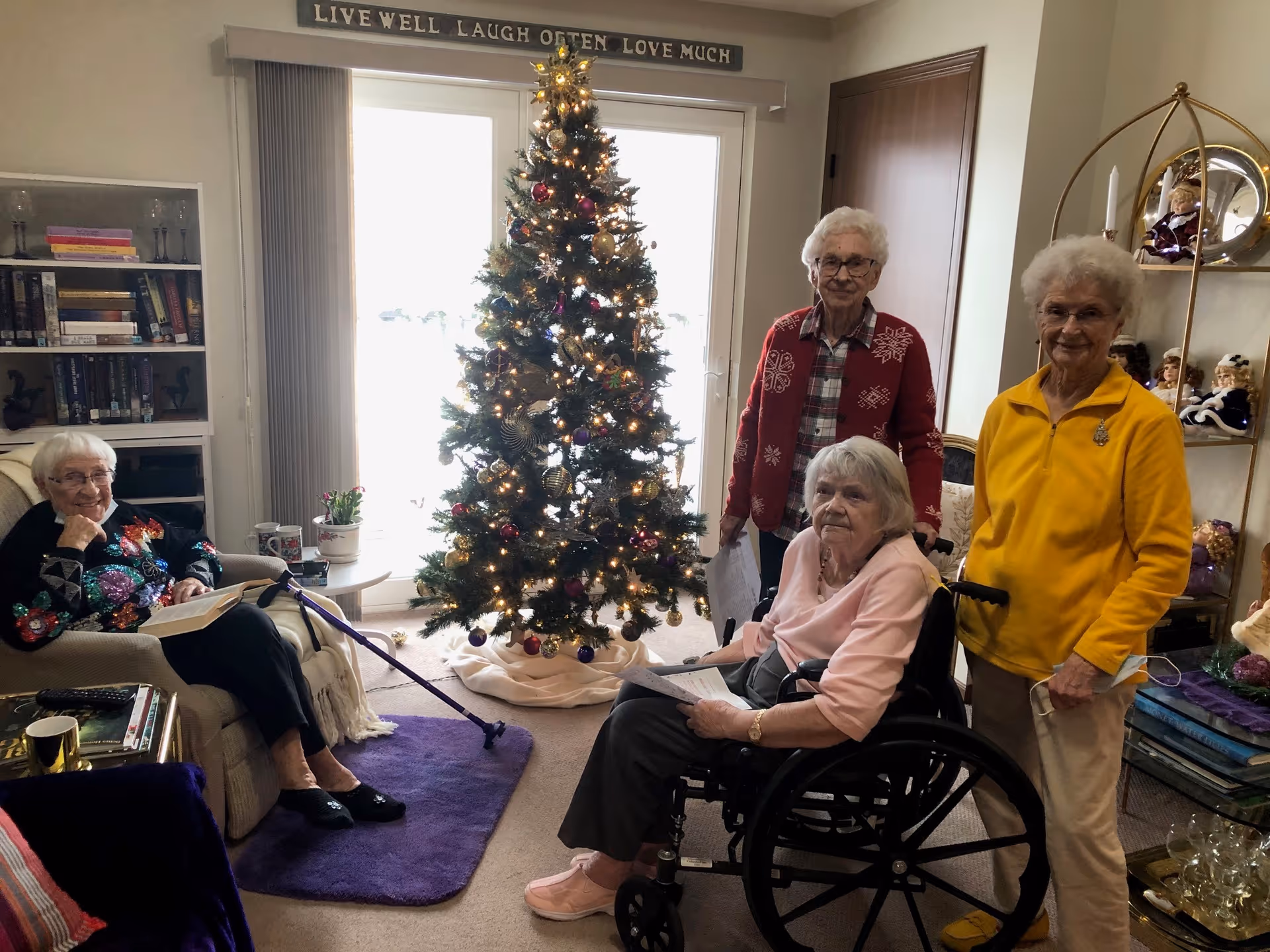 Four elderly women gathered in a cozy living room decorated for Christmas with a lit Christmas tree in the center. One woman is seated in a wheelchair holding papers, another is sitting on a chair reading a book, and two women are standing behind the wheelchair. The room has a bookshelf, a small table with plants, and a decorative shelf with dolls. A sign above the window reads 'LIVE WELL LAUGH OFTEN LOVE MUCH'.