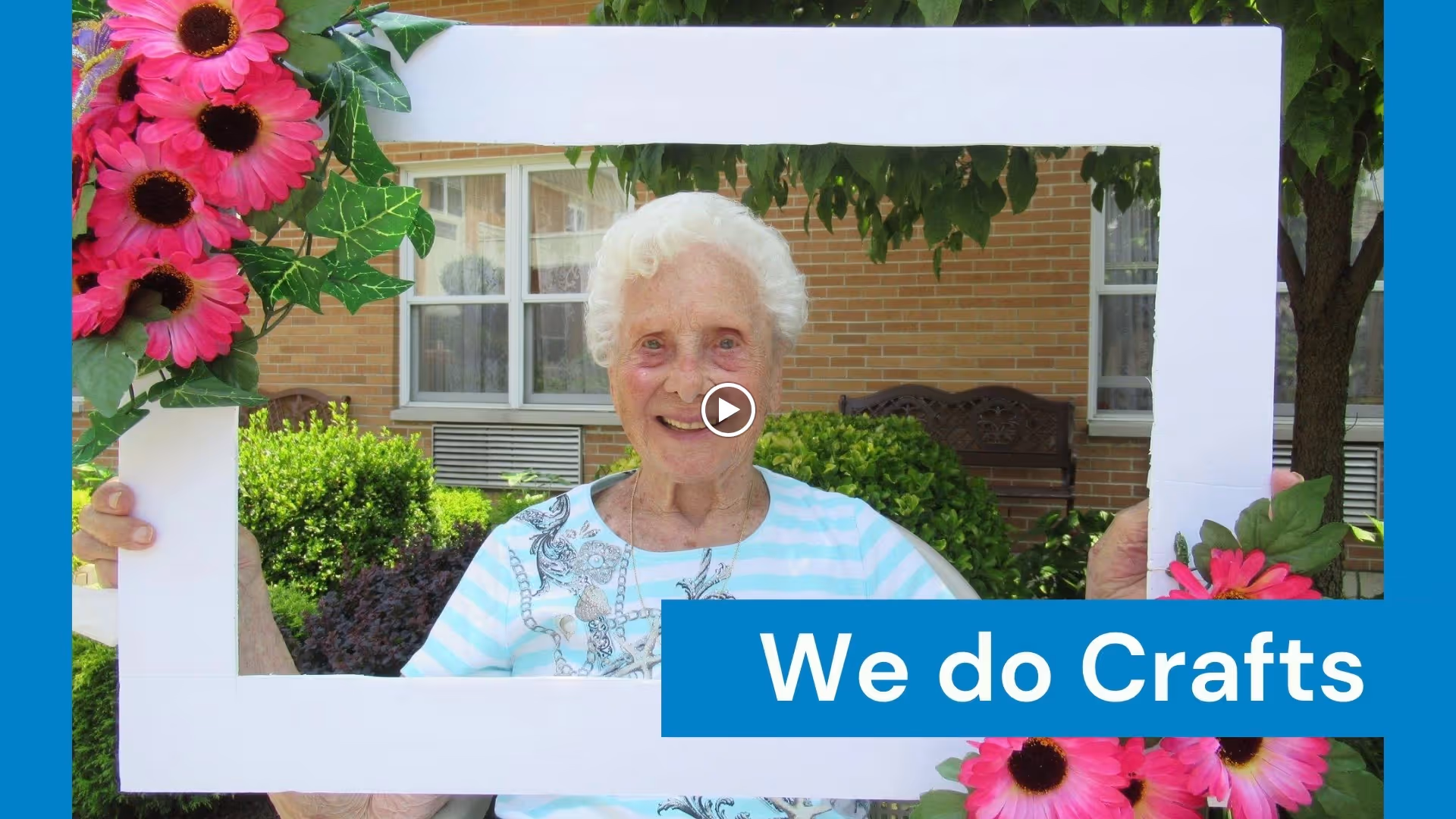 An elderly woman with white hair smiling and holding a large white rectangular frame decorated with pink flowers and green leaves. She is outdoors with greenery and a brick building in the background. A blue banner with the text 'We do Crafts' is partially covering the bottom right of the frame.