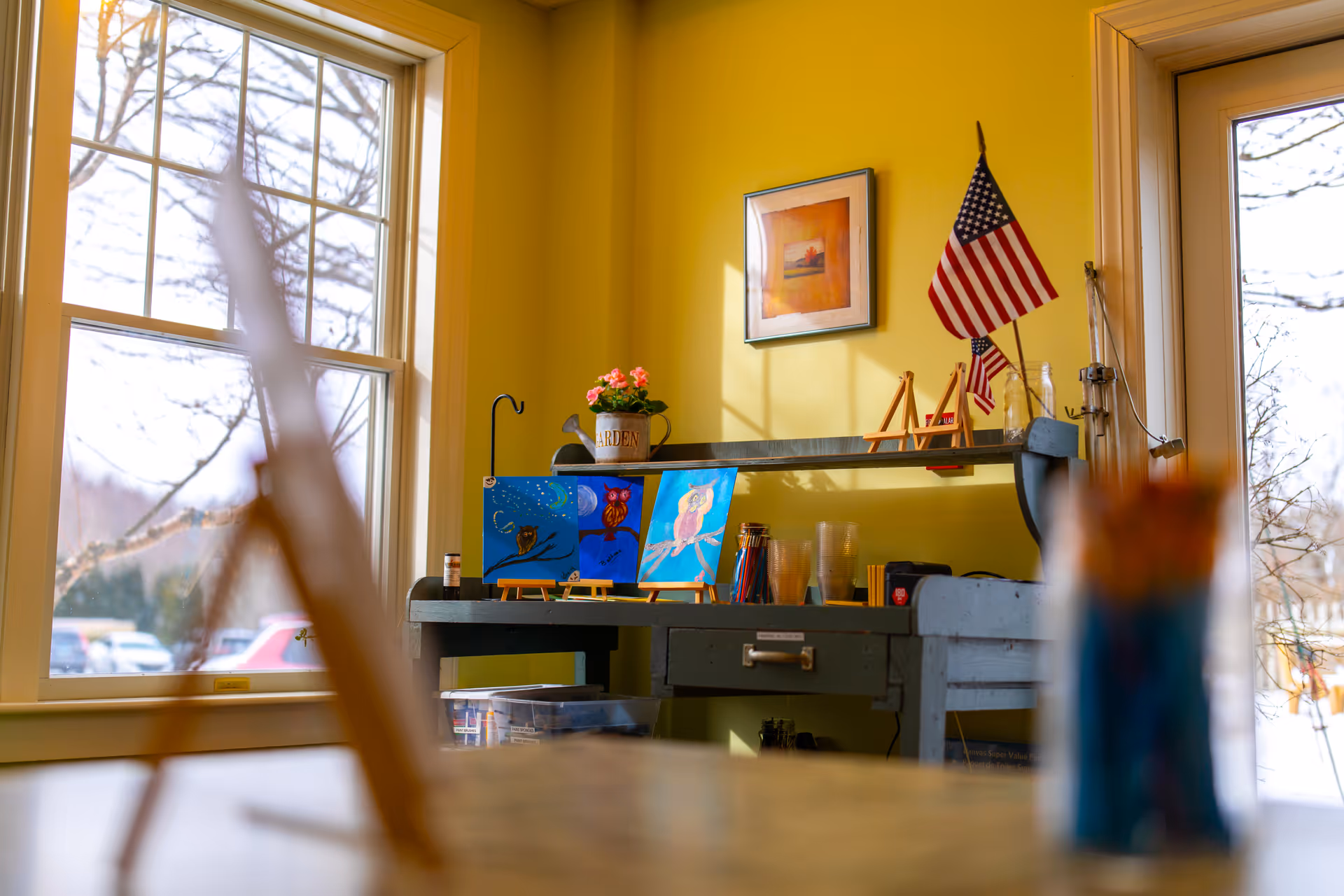 A bright room with yellow walls featuring a desk with small paintings of owls on easels, an American flag, a potted plant, and art supplies. Large windows let in natural light and show a view of trees and parked cars outside.