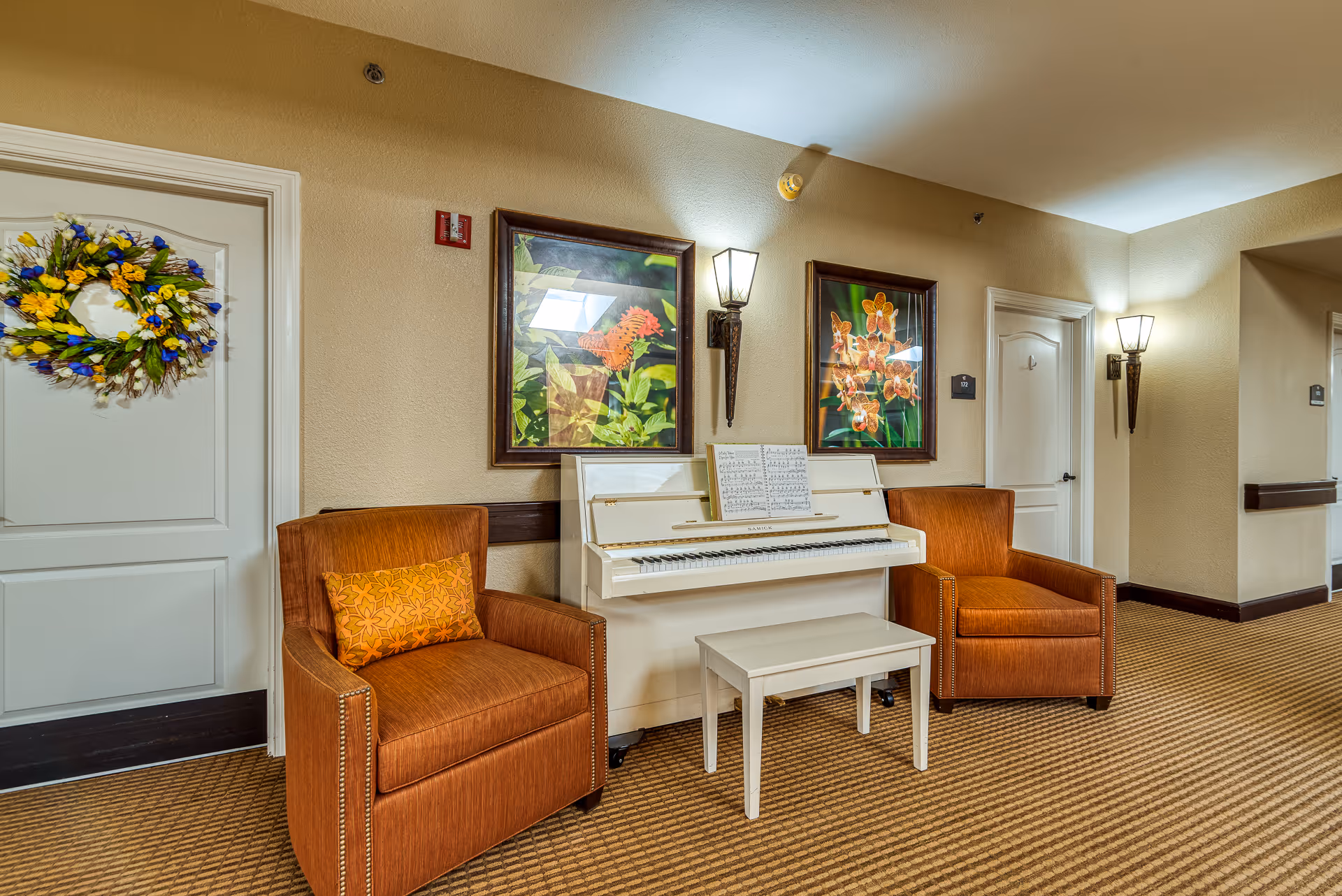 A hallway seating area with two orange armchairs flanking a white upright piano beneath framed floral artwork.