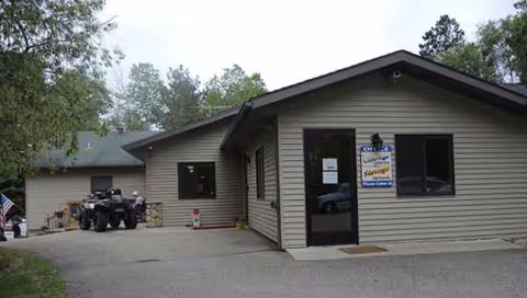 Single-story beige building exterior with front entrance and parked vehicles nearby.