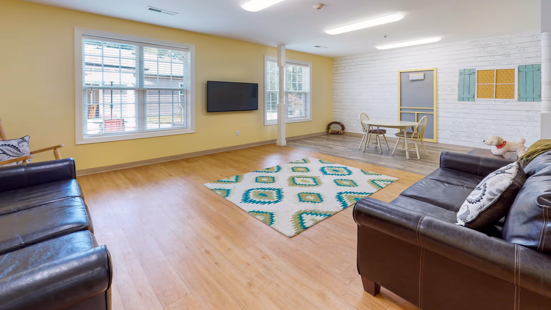 Spacious bright common living room with leather sofas, an area rug, a wall-mounted TV, and a small table near windows.