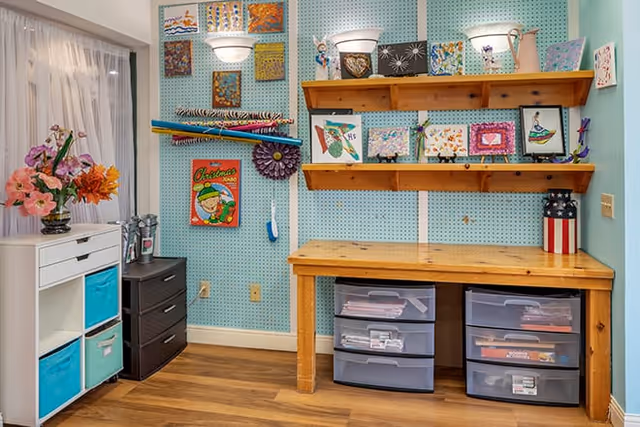 A craft room with a wooden table and two plastic drawer units underneath. The wall behind the table is light blue with pegboard panels displaying various colorful artwork and decorations. Two wooden shelves hold more art pieces and decorative items. To the left, there is a white storage unit with blue fabric bins and a vase of flowers on top. The floor is wooden, and a window with sheer white curtains is partially visible on the left side.