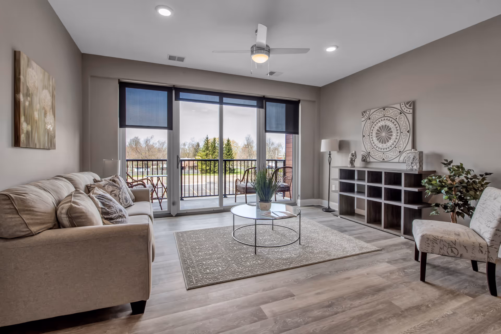 A modern living room with a beige sofa adorned with patterned pillows on the left, a round glass coffee table on a textured rug in the center, and a dark wooden shelving unit with decorative items on the right. A patterned armchair and a potted plant are also visible. Large sliding glass doors with blue roller shades open to a balcony with outdoor chairs and a view of trees and a parking lot. The room has light wood flooring, neutral-colored walls, recessed ceiling lights, and a ceiling fan with a light.