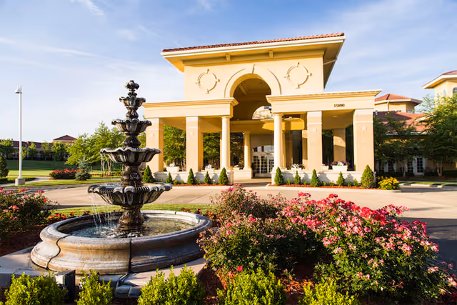 Front exterior view of a senior living facility named Santa Marta with a large covered entrance supported by columns, a multi-tiered water fountain in the foreground, and landscaped bushes and flowers around the driveway.