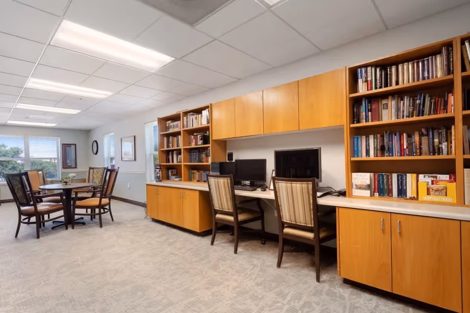 A well-lit room with a carpeted floor featuring a wooden table with four chairs near windows. Along one wall, there are built-in wooden shelves filled with books and two computer workstations with chairs. The room has a calm and organized atmosphere.