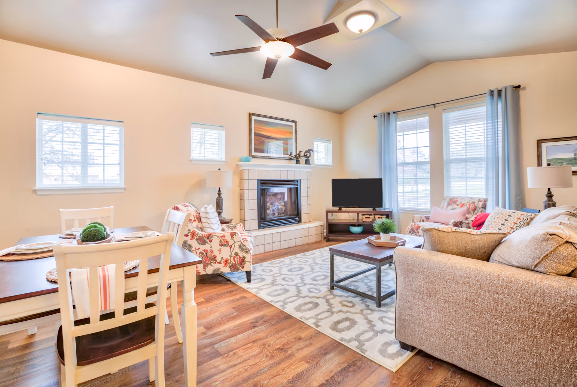 A bright and cozy living room with a beige sofa, floral armchair, wooden coffee table on a patterned rug, and a fireplace with a framed picture above it. There is a ceiling fan with lights, large windows with light blue curtains, and a dining table with white chairs set for four people.