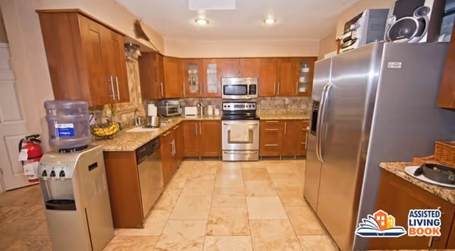 A spacious kitchen with wooden cabinets, granite countertops, stainless steel appliances including a refrigerator, oven, microwave, and dishwasher. There is a water dispenser and a fire extinguisher on the left side. The floor is tiled with large beige tiles.