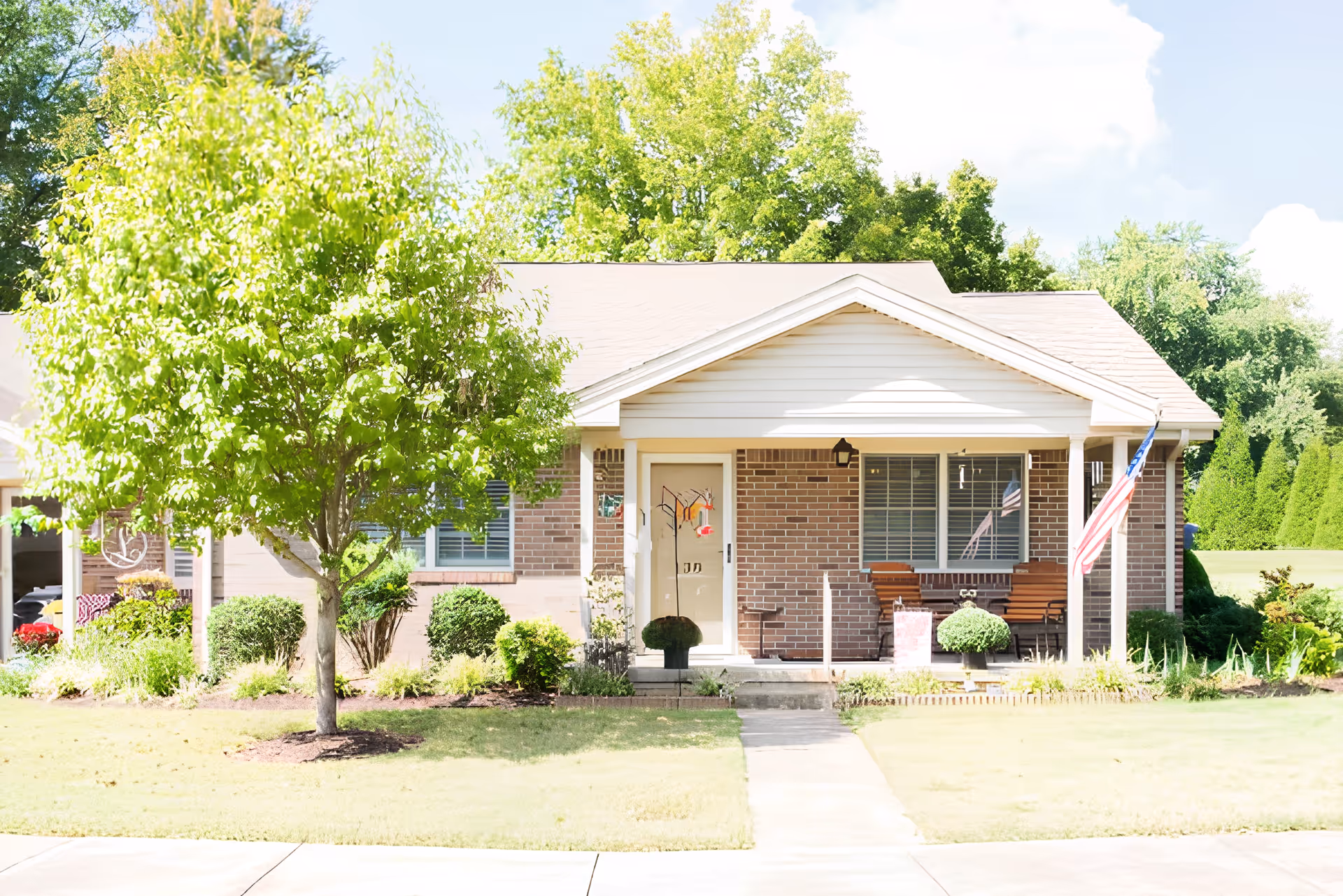 Front exterior view of a single-story brick building with a small porch, a bench, an American flag, and a tree in the front yard under a clear sky.