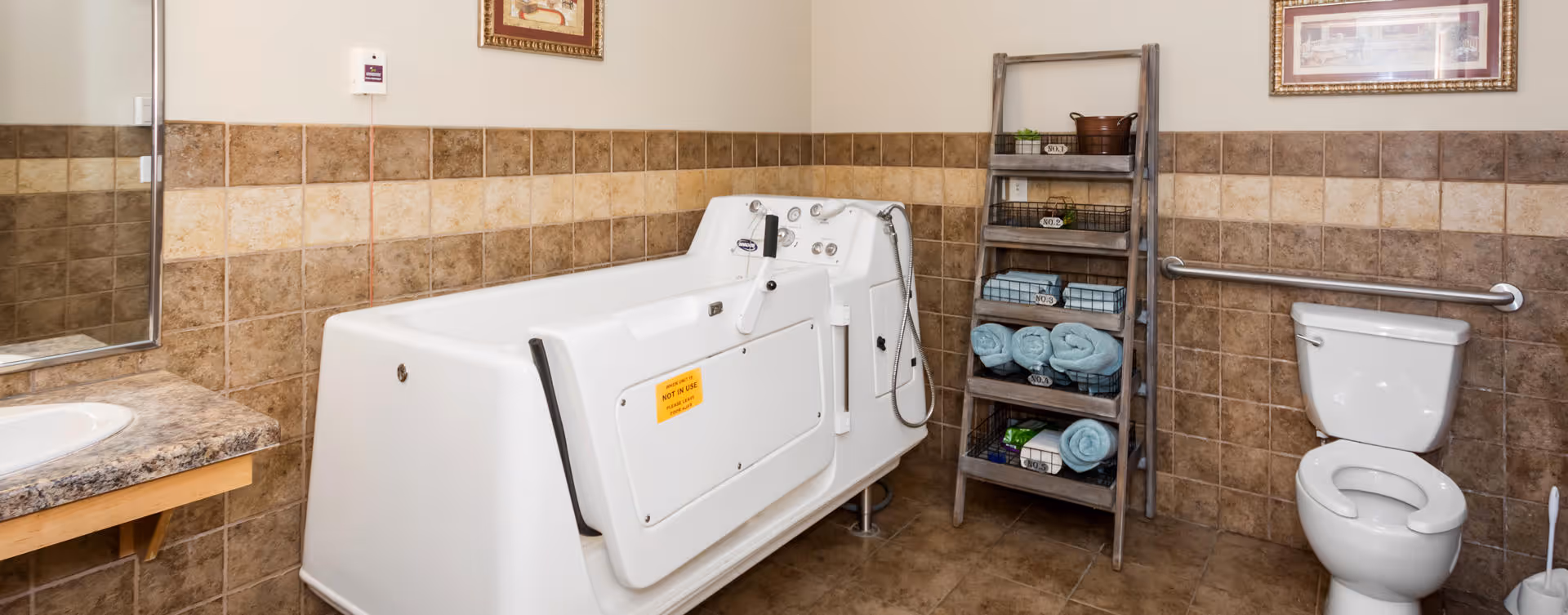 A bathroom in an assisted living facility featuring a walk-in bathtub with safety handles, a toilet with a grab bar, a wooden ladder shelf holding neatly rolled towels and baskets, a sink with a granite countertop, and tiled walls and floor.