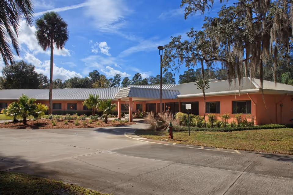 Exterior view of Wellsprings Residence LLC, a single-story building with a light gray metal roof and peach-colored walls, surrounded by landscaped greenery including palm trees and shrubs under a blue sky with scattered clouds.