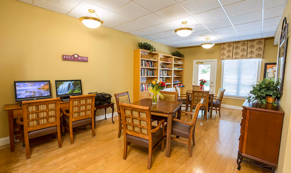A cozy common area with wooden tables and chairs, a bookshelf filled with books, two computer monitors on a desk, and a window with blinds. The walls are painted yellow, and there are ceiling lights providing warm illumination. A small sign on the wall reads 'Paris Bistro'.