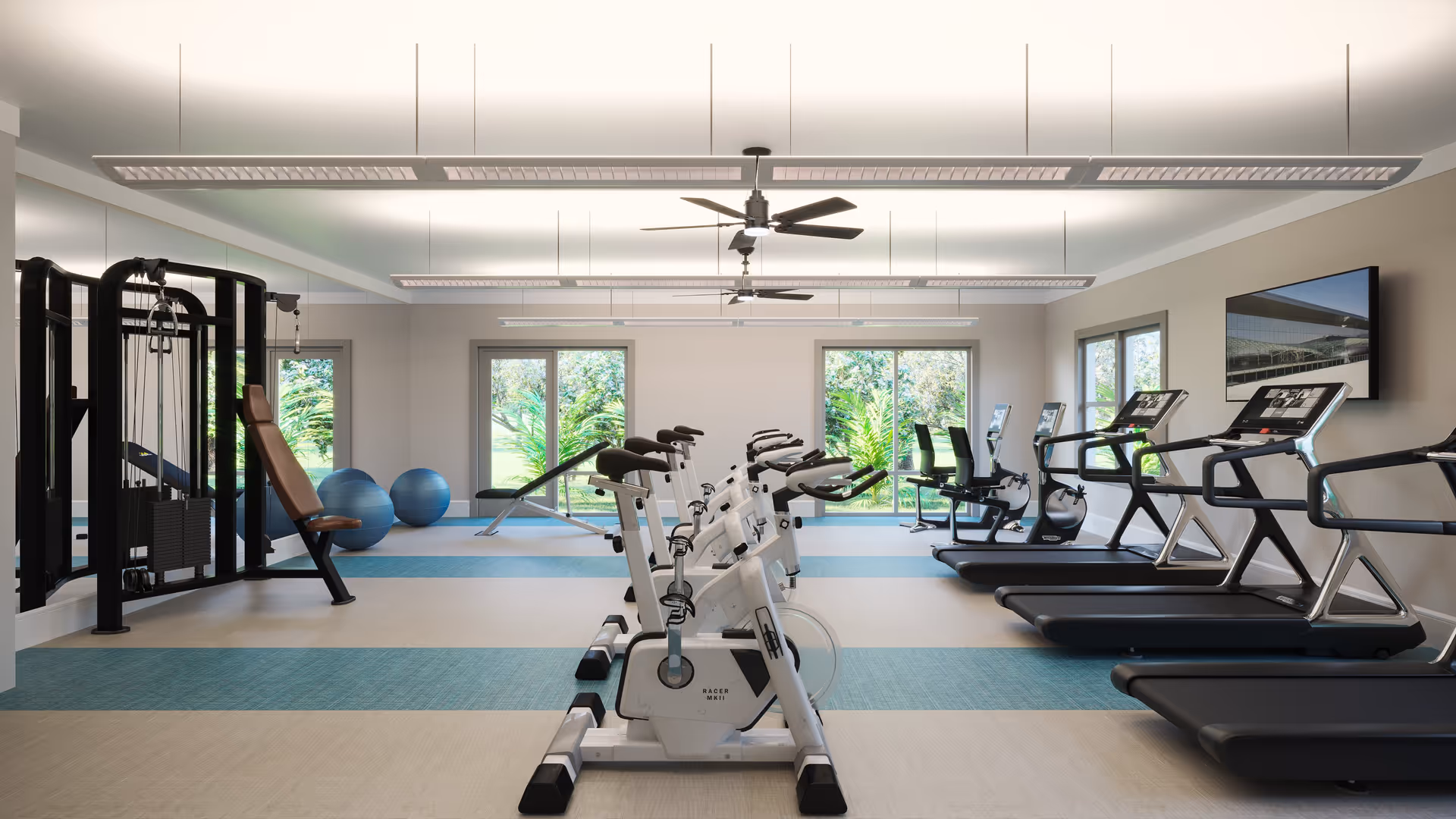 A bright and spacious fitness room with exercise bikes, treadmills, weight machines, and exercise balls. Large windows provide a view of greenery outside, and ceiling fans and modern lighting fixtures are installed overhead.