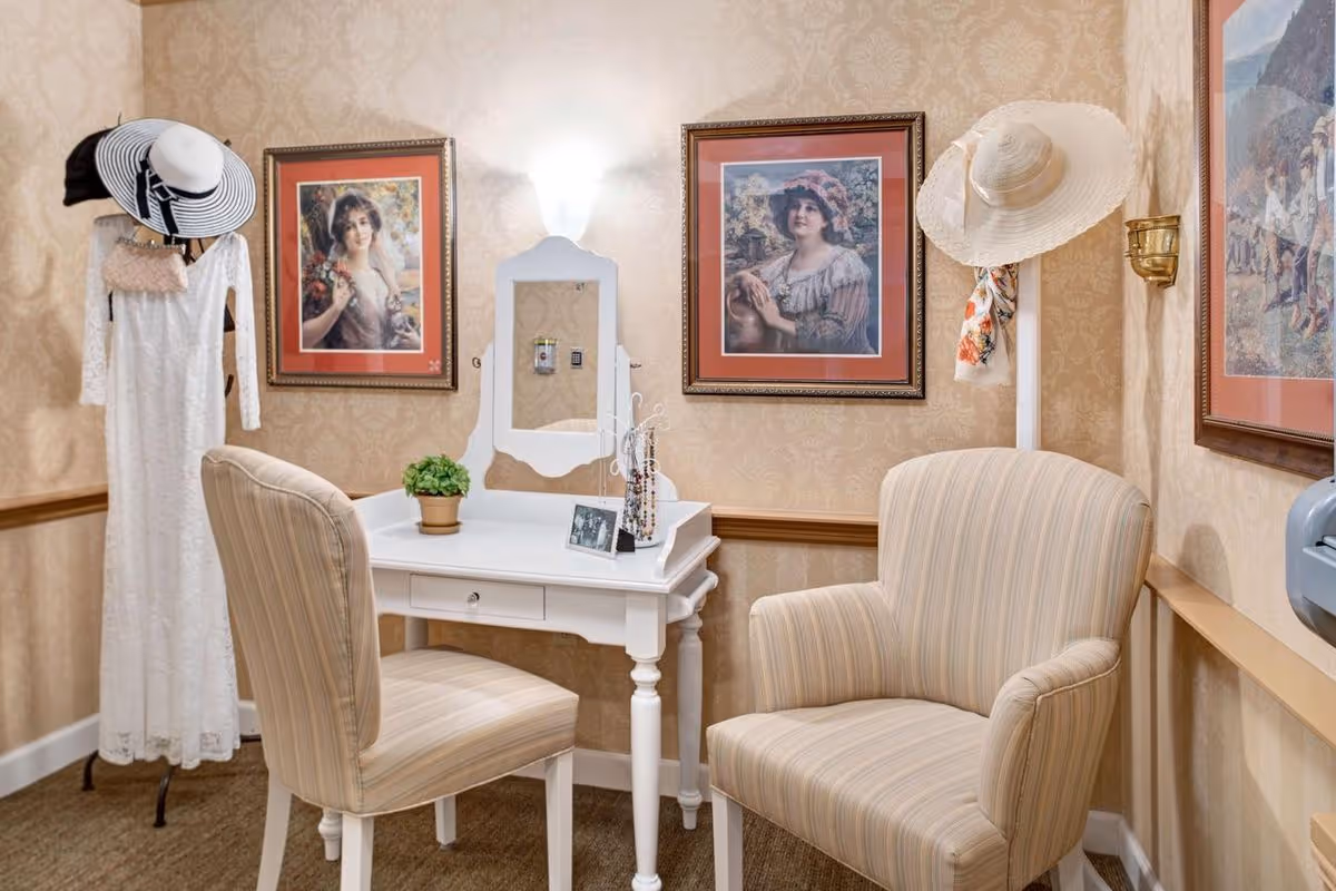 A cozy interior corner with a white vanity table and mirror, a striped upholstered chair, and an armchair. The walls are decorated with framed vintage-style portraits and patterned wallpaper. A coat rack holds a white dress, hats, and a small purse.
