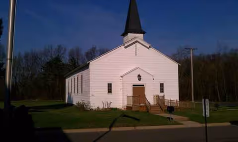 A white church with a tall black steeple sits on a grassy area.