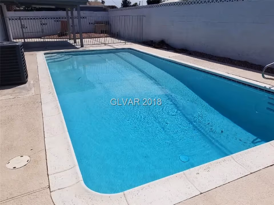 Outdoor swimming pool with clear blue water surrounded by a concrete deck and enclosed by a white fence with a gate.