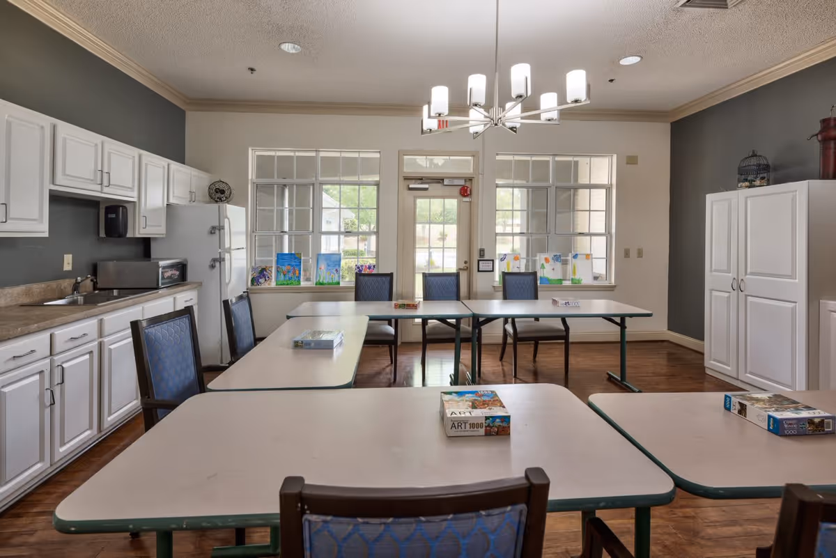 A well-lit activity room with several tables and chairs arranged for group activities. The room features white cabinets and a countertop with a sink on the left side, a refrigerator, and windows with colorful artwork displayed on the sills. A modern chandelier hangs from the ceiling, and the floor is wooden.