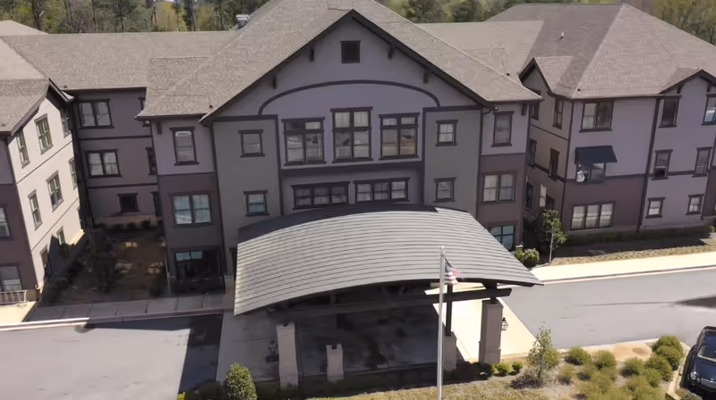 Front exterior view of The Phoenix at Milton senior living facility showing a three-story building with multiple windows, a covered entrance with a curved roof, an American flag on a pole, and surrounding landscaping.