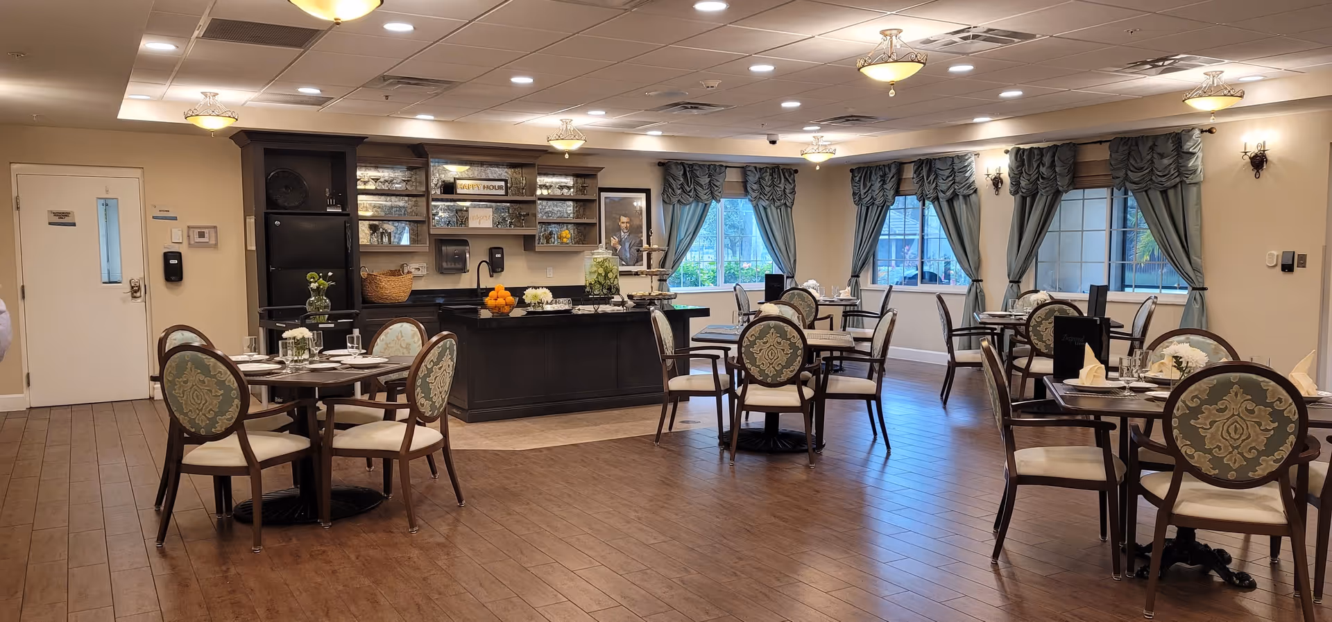 A spacious dining room with several round tables set with plates, glasses, and napkins. The room features wooden flooring, large windows with blue curtains, and a central serving area with shelves holding glassware and decorative items. The lighting is warm with ceiling fixtures and wall sconces.