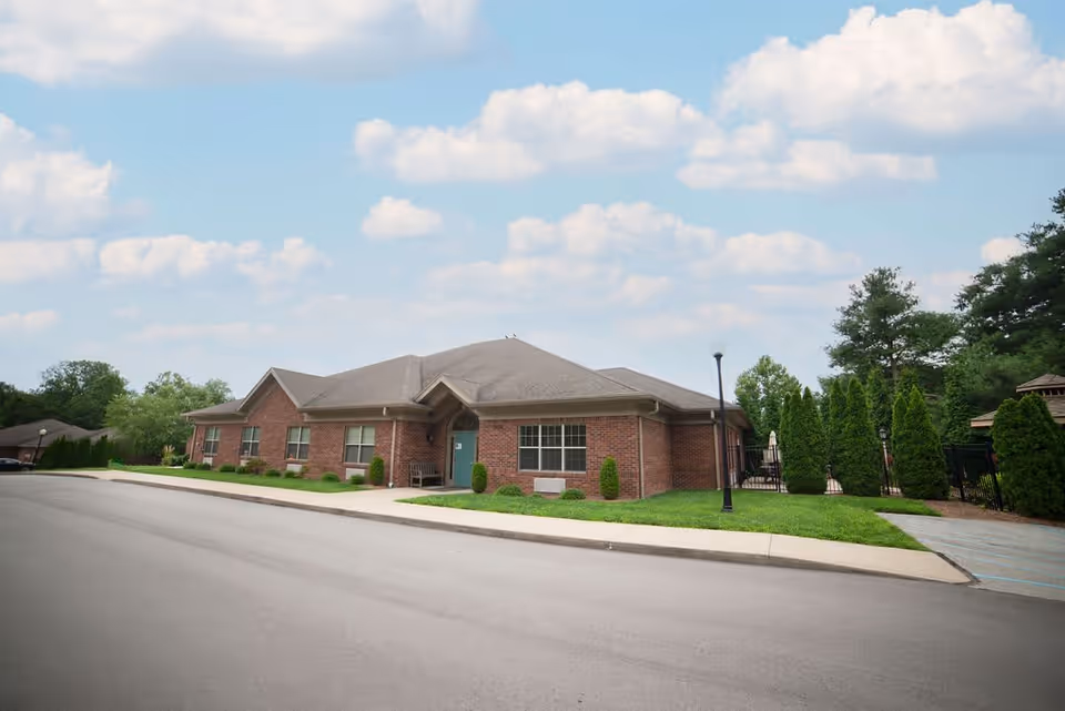 Exterior view of a single-story brick building with a gray roof, surrounded by green grass, shrubs, and trees under a partly cloudy sky. A paved road runs in front of the building, and there is a sidewalk with a lamppost near the entrance.