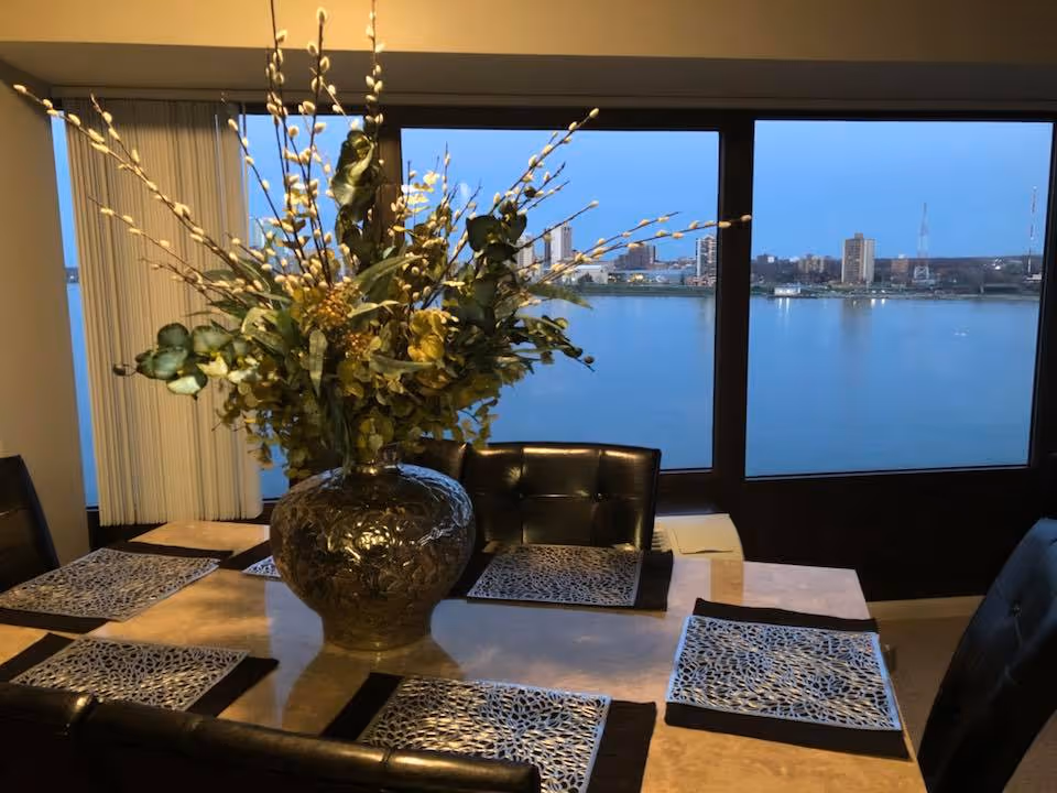 Dining table with a large floral centerpiece and placemats in front of floor-to-ceiling windows overlooking a river and city skyline.