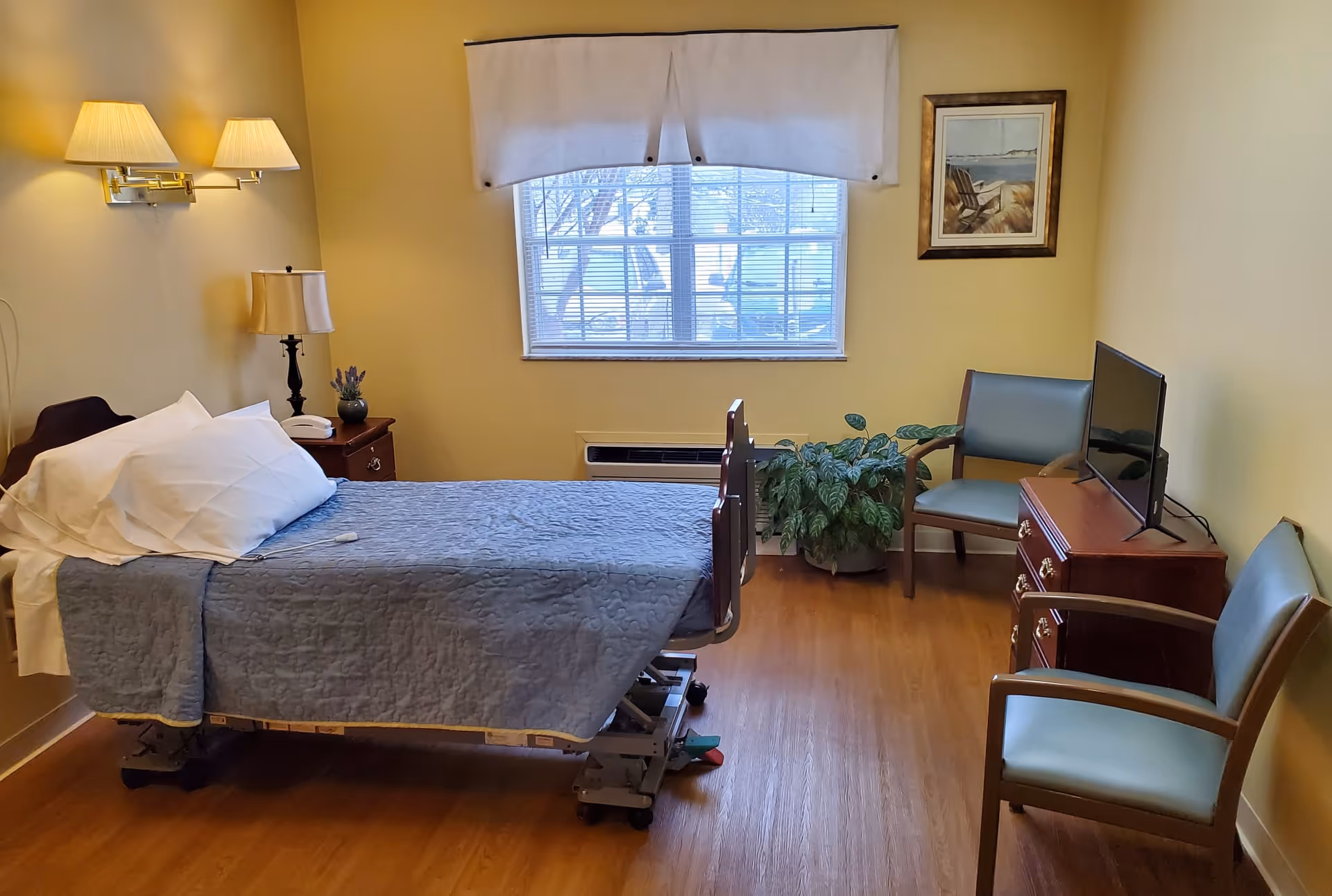 A senior care facility bedroom with a single hospital-style bed covered with a blue quilt and white pillows. The room has wooden flooring, two green chairs, a wooden dresser with a flat-screen TV, a nightstand with a lamp and a small plant, a window with blinds and a white valance, and a framed picture on the wall.