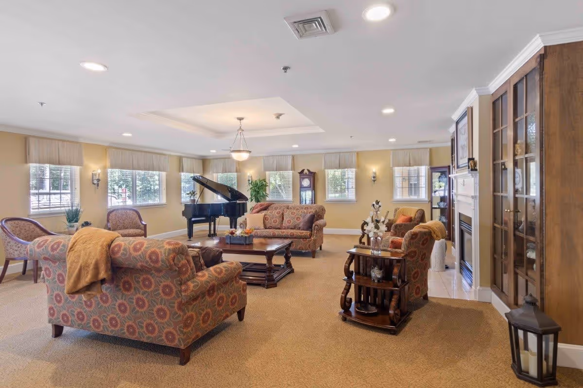 A spacious and well-lit living room with patterned upholstered sofas and armchairs arranged around a wooden coffee table. The room features large windows with beige curtains, a grand piano, a grandfather clock, a fireplace, and wooden display cabinets. The carpet is beige, and the walls are painted a soft yellow.