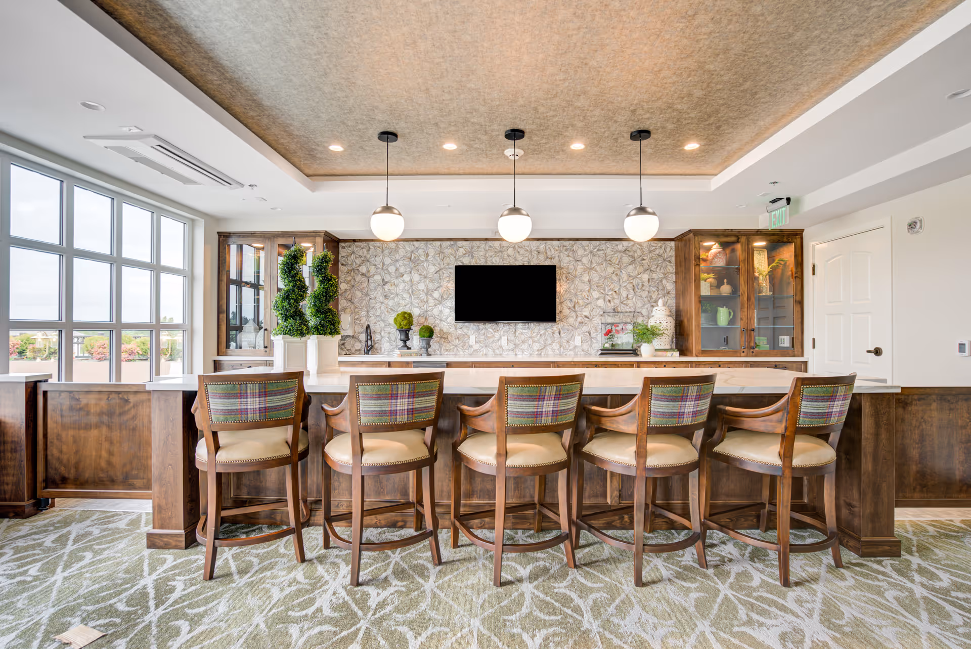 A modern and elegant interior space featuring a long wooden counter with six high-back chairs upholstered with plaid fabric on the backrests and beige seats. Behind the counter is a decorative wall with a mounted flat-screen TV, flanked by wooden cabinets with glass doors displaying decorative items. Three round pendant lights hang from the ceiling above the counter. Large windows on the left side allow natural light to fill the room, and the floor is covered with a patterned carpet.