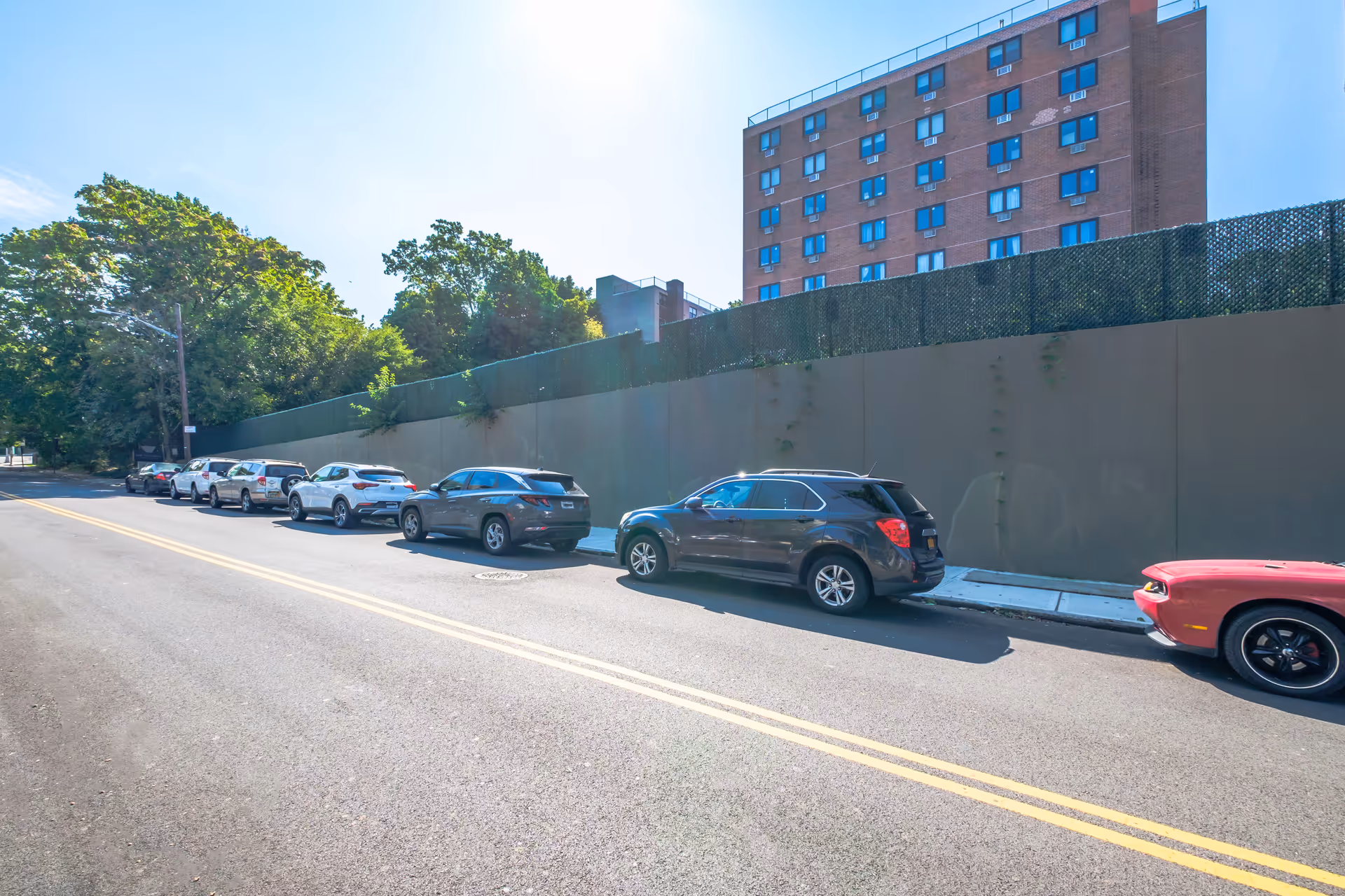 Cars parked along a city street beside a tall fence with a multi-story brick building and trees in the background.