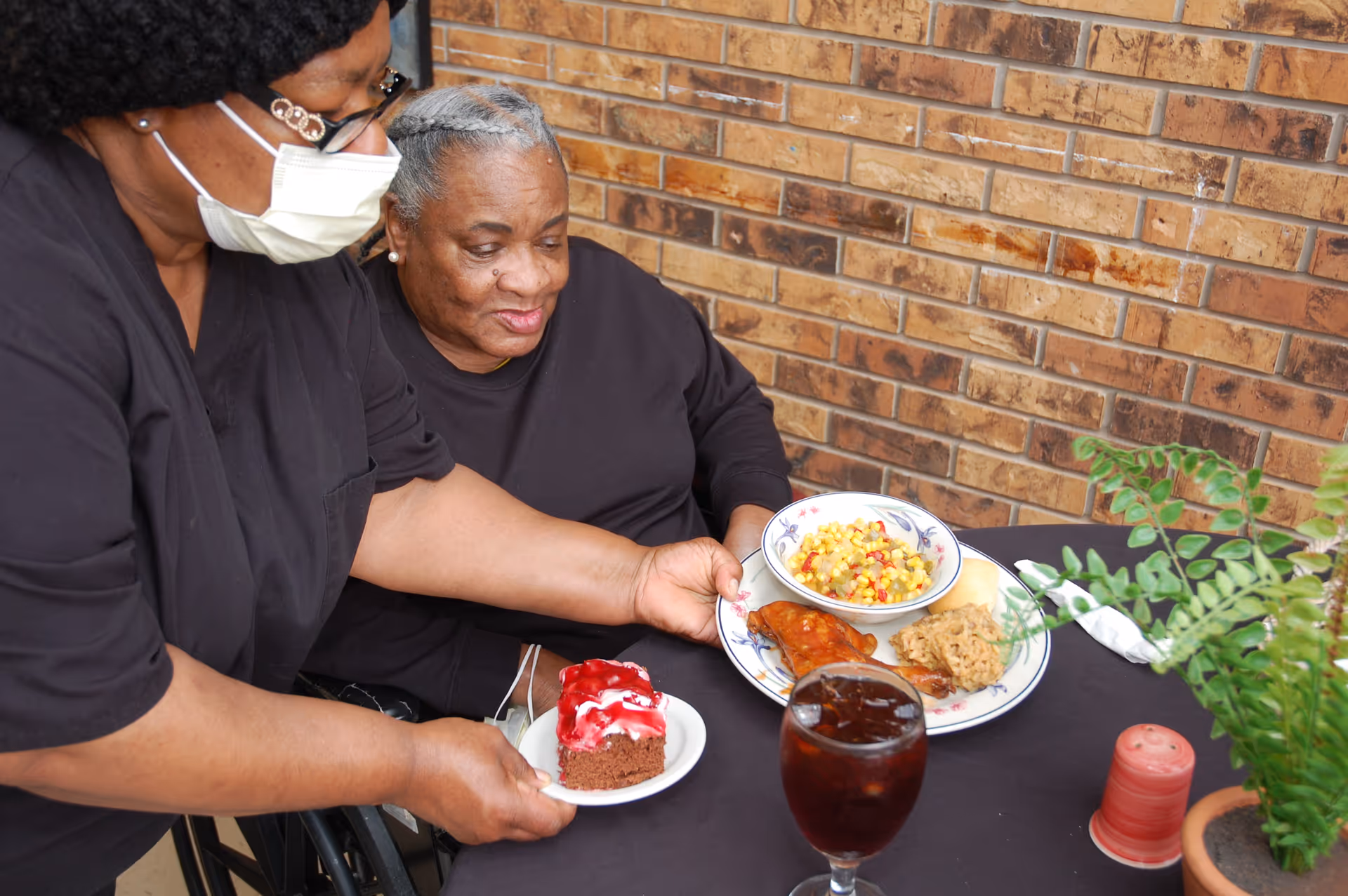 A caregiver wearing a mask serves a plate of food and a dessert to an elderly woman seated at a table with a brick wall background. The table has a glass of iced tea, a potted plant, and a salt shaker.