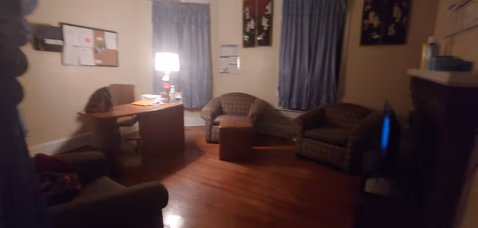 A dimly lit room with wooden flooring featuring a desk with a lamp, two armchairs, a small ottoman, a bulletin board on the wall, and two windows with blue curtains. There is also a fireplace on the right side of the room.